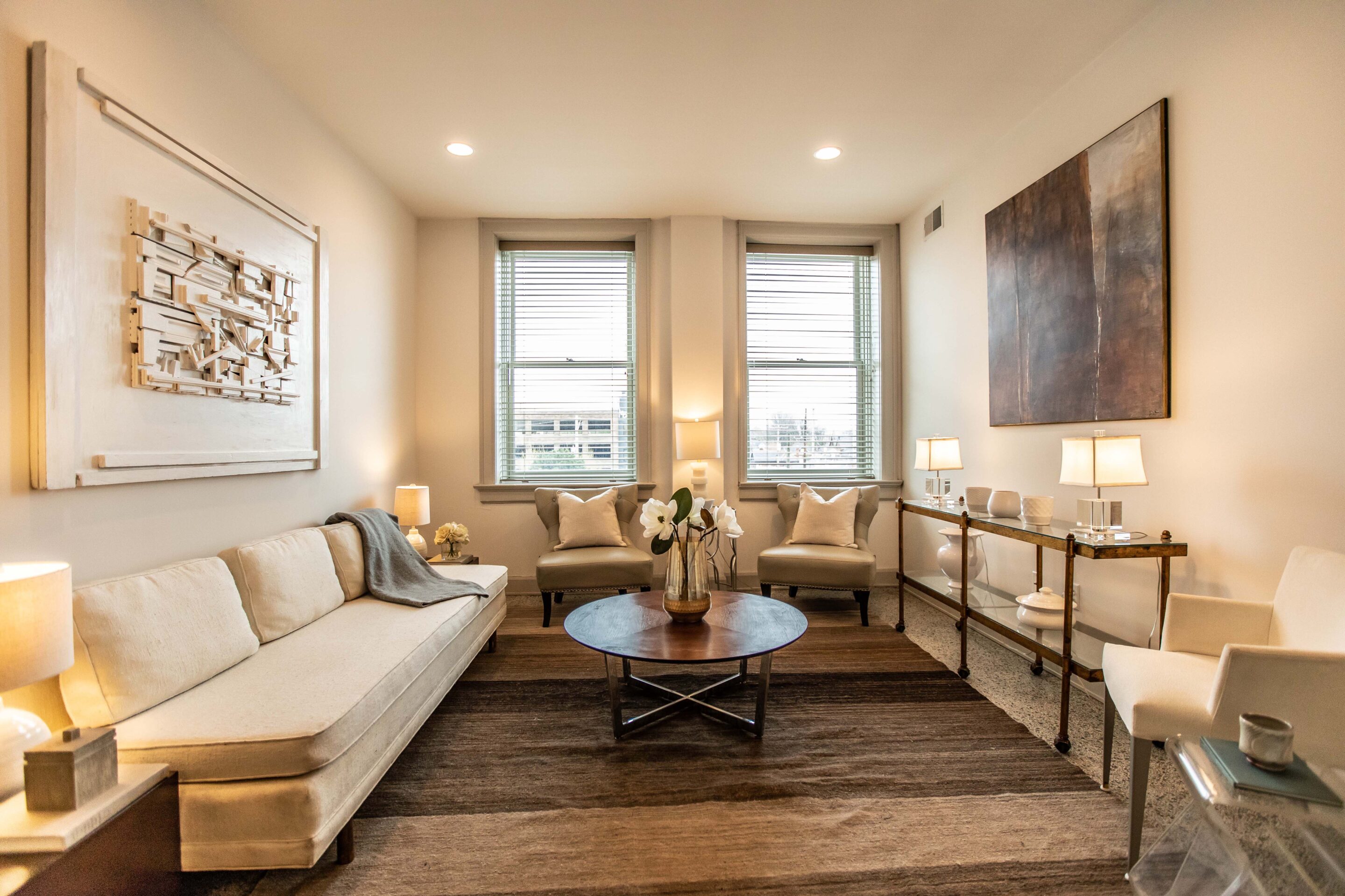 A modern living room with a cream sofa, two windows, abstract wall art, a round coffee table, and neutral decor.