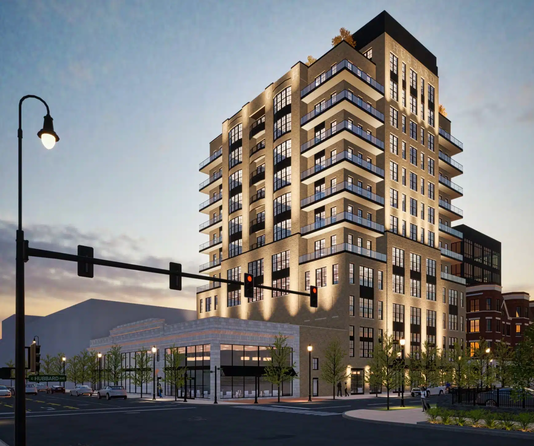 Modern multi-story building with illuminated windows at dusk, featuring commercial spaces on the ground floor and a street intersection with traffic lights in the foreground.