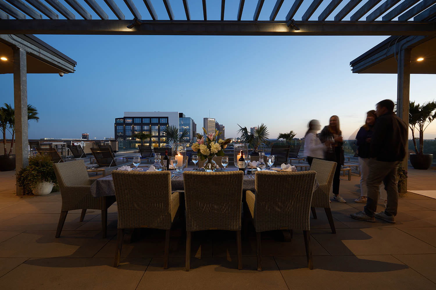 Outdoor dining setup on a terrace at dusk with a table, chairs, flowers, and candles. Four people stand nearby under a pergola. Buildings and trees are in the background.