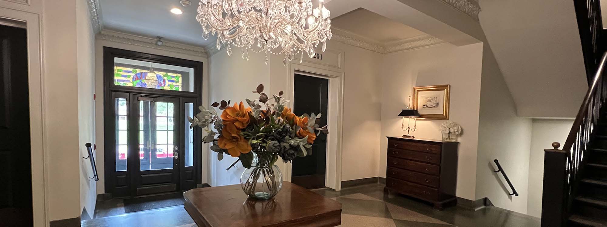 Interior of a foyer with a chandelier, a vase of orange flowers on a wooden table, a staircase, a painting on the wall, and double doors with stained glass.