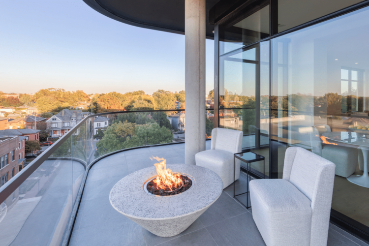 A modern balcony with a glass barrier features a circular fire pit, two white chairs, and a view of trees and houses in the distance.