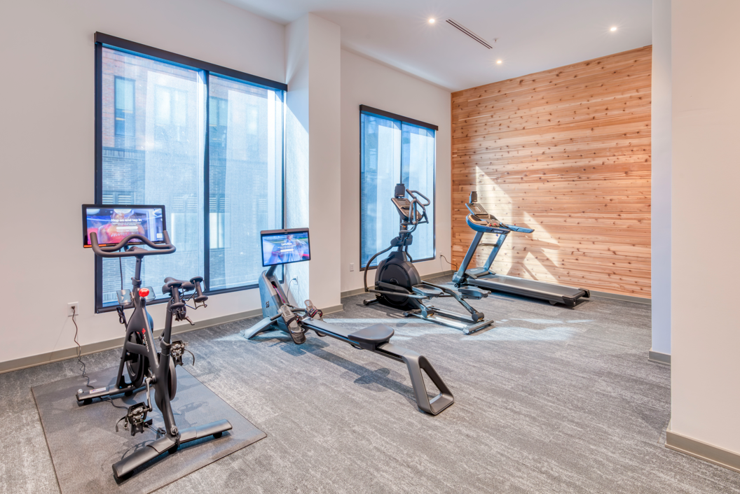 A gym with an exercise bike, rowing machine, elliptical, and treadmill. The room has large windows and a wood-paneled wall.