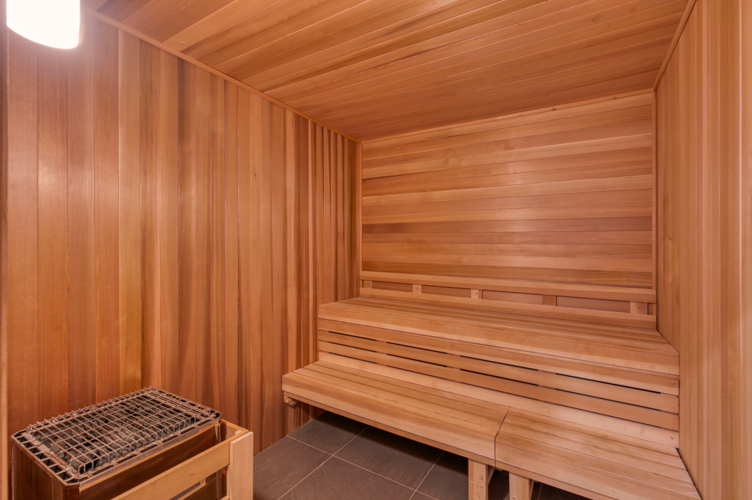 Interior of a wooden sauna with a multi-level bench and a covered heater on the left. A light fixture is mounted on the wall.