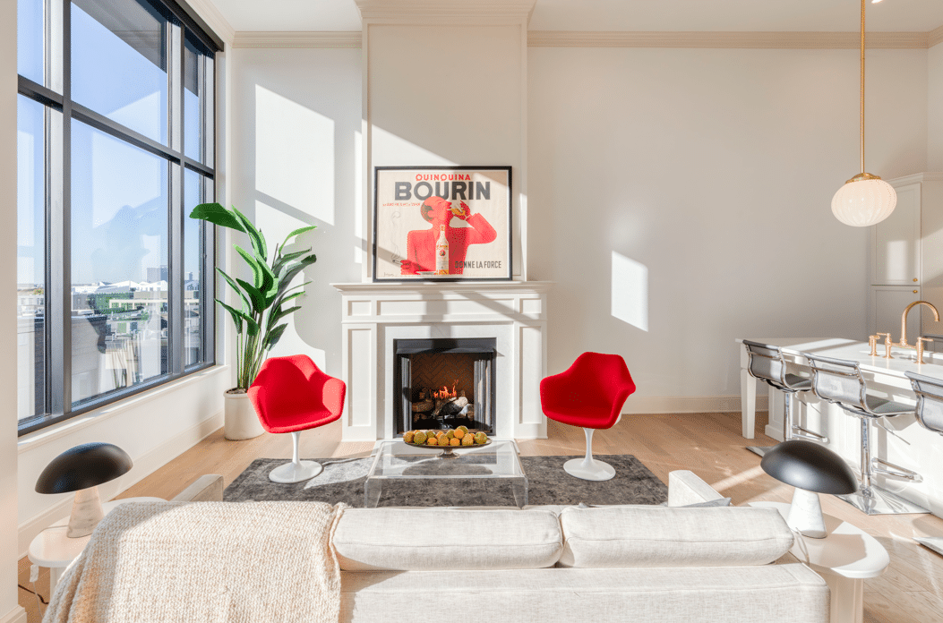 Modern living room with two red chairs facing a fireplace. Large windows allow natural light. A poster and a plant decorate the space. A kitchen island with stools is visible to the right.