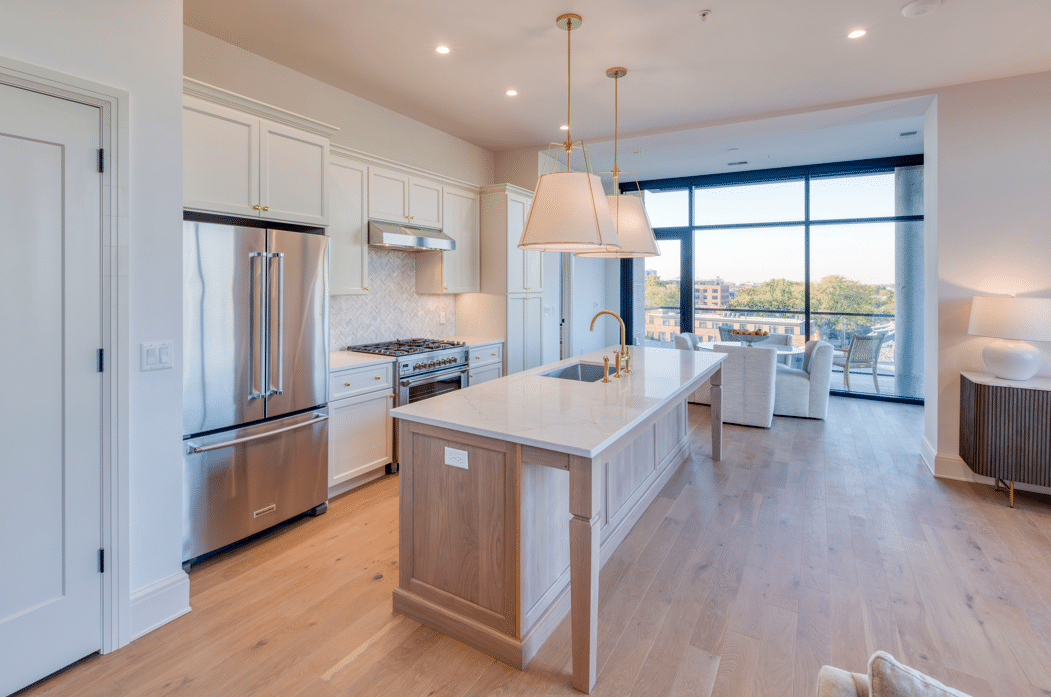 Modern kitchen with a long central island, stainless steel appliances, pendant lighting, and large windows overlooking a city view.