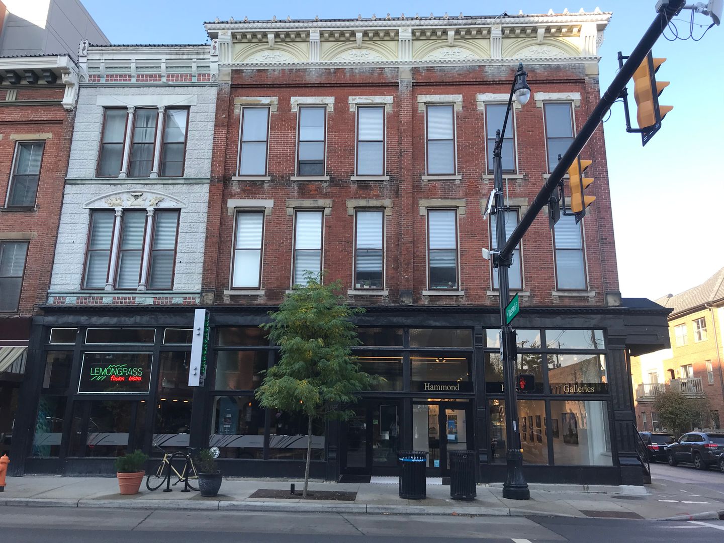 A three-story brick building with ground floor businesses, including a restaurant and bookstore. The facade features large windows. A tree and a bicycle are in front. Traffic lights are visible.
