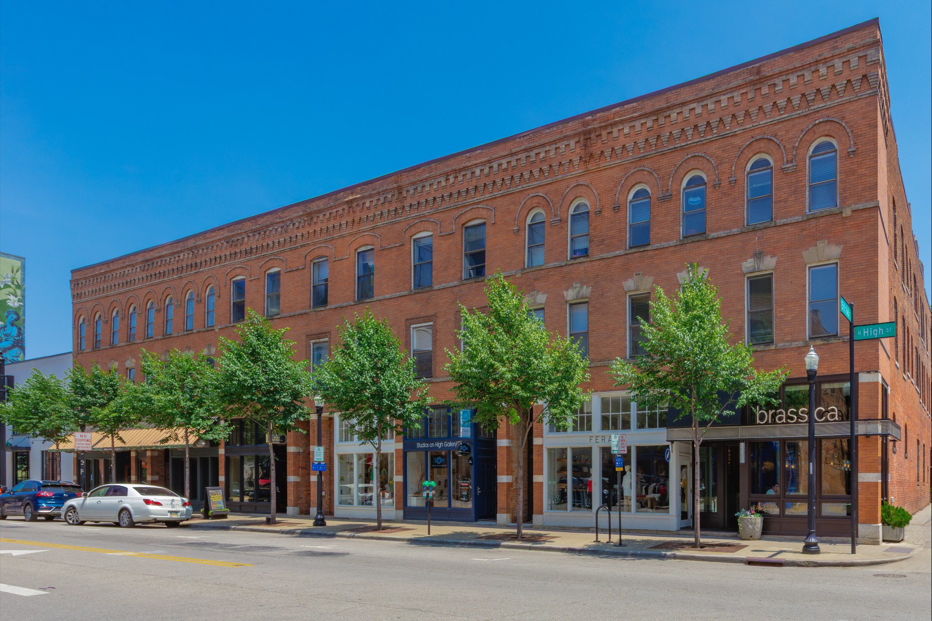 A three-story red brick building with storefronts and trees lining the sidewalk; parked cars are visible on the street.